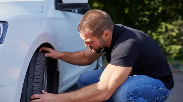 Man checking the alignment or condition of a car tire on a white vehicle, crouched down and focused on the front wheel.