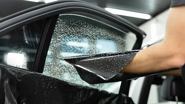 Close-up of a person applying tinted film to a car window, with water droplets visible on the glass to aid adhesion during installation.