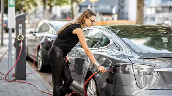 Woman charging a gray electric car at a public charging station on a city street, holding a smartphone in one hand while plugging in the charging cable.